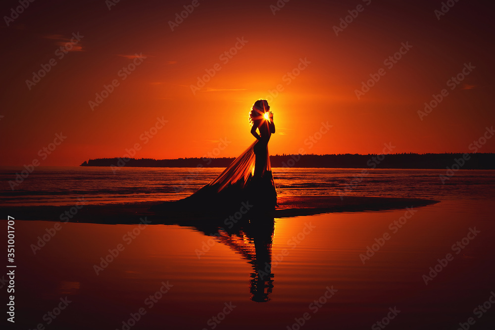 Silhouette of a young girl in a long transparent dress on the beach against the sunset sky.