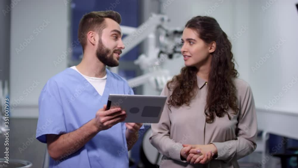 Handheld medium shot of cheerful male dentist in scrubs holding tablet and showing something to female patient during consultation in dental clinic Handheld medium shot of cheerful male dentist in scrubs holding tablet and showing something to female patient during consultation in dental clinic