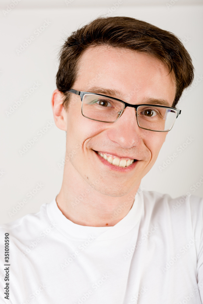 Young smiley guy with dark hair, glasses and a T-shirt making selfies on a white wall.