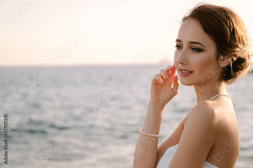 Portrait of beautiful young smiling girl in the white dress in greek style walking along the sea promenade. Summer, sunset time. Bride, wedding at sea 