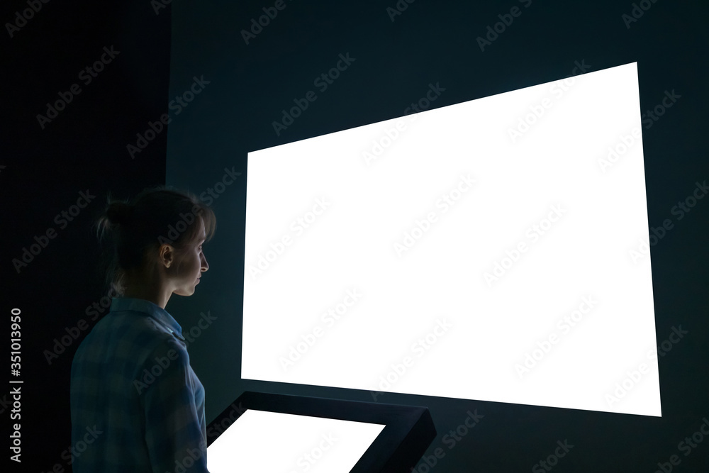 Woman looking at white blank large interactive wall display in dark room of modern technology exhibition. Evening time, low light. Mock up, template, copyspace, education and technology concept Woman looking at white blank large interactive wall display in dark room of modern technology exhibition. Evening time, low light. Mock up, template, copyspace, education and technology concept