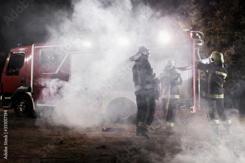 Team of professional firefighters with water hose in front of a firetruck with smoke in the air.