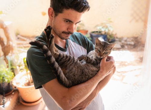 Fotografie Hombre joven sostiene en sus brazos a un gato atigrado de color gris y mira haci