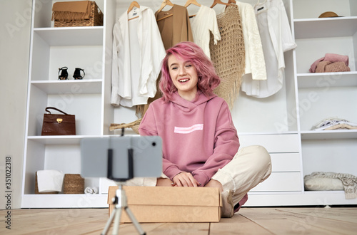 Happy hipster gen z teen girl fashion social media channel blogger with pink hair wear hoodie recording unboxing vlog on phone video camera tutorial sit on floor with online store clothes order box. photo