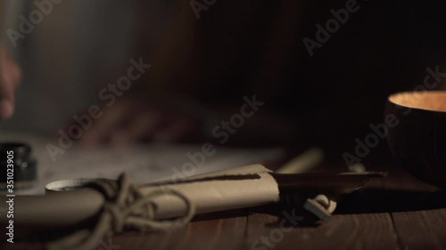 A medieval man uses an old pen with ink to write on an a paper lit by a candle,slow motion