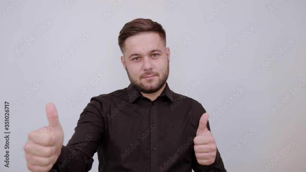 Young Bearded Dark Haired Man In Black Stylish Shirt On Gray Background, Serious Male Holds Thumb Finger Up, Looks Straight Ahead. The Concept Of Cool People, Leaders Young Bearded Dark Haired Man In Black Stylish Shirt On Gray Background, Serious Male Holds Thumb Finger Up, Looks Straight Ahead. The Concept Of Cool People, Leaders