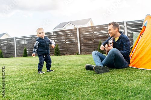 Toddler son and his father have fun in the backyard. Dad and son are camped on the lawn, daddy plays guitar, baby boy dances and laughs