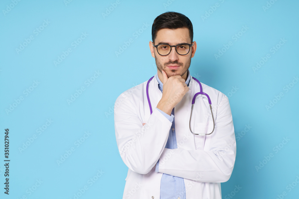 Male doctor holding his chin, thinking about difficult medical case, feeling doubt about cure, wearing white robe and glasses, isolated on blue background Male doctor holding his chin, thinking about difficult medical case, feeling doubt about cure, wearing white robe and glasses, isolated on blue background