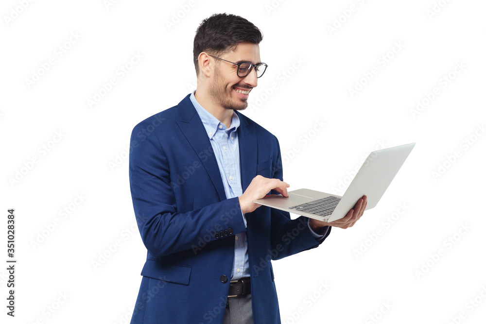 Smiling young man standing in blue suit and glasses, holding open laptop, watching funny content, feeling happy and relaxed, isolated on white background