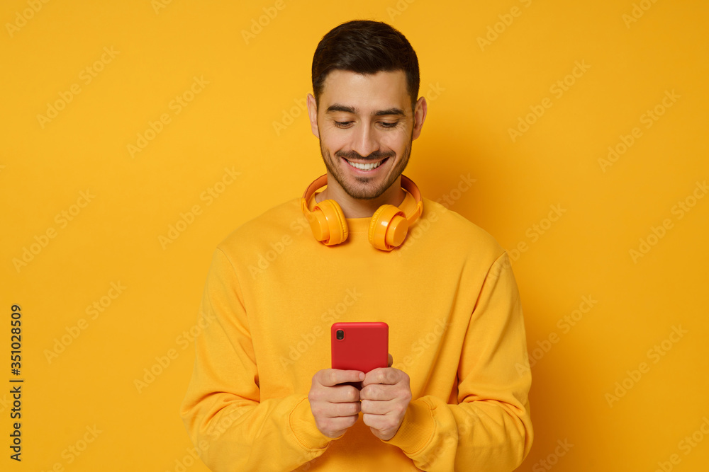 Young trendy man wearing sweatshirt and wireless headphones, smiling while looking at content on phone screen, isolated on yellow background Young trendy man wearing sweatshirt and wireless headphones, smiling while looking at content on phone screen, isolated on yellow background