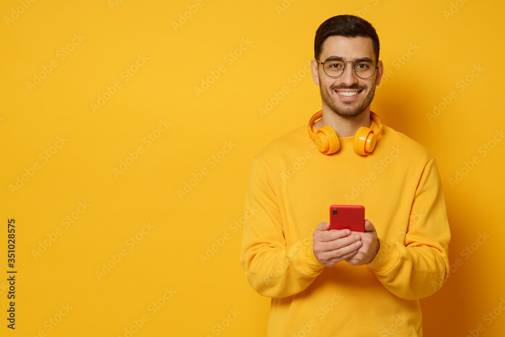 Banner of young man holding phone in hands, wearing wireless headphones, sweatshirt and glasses, smiling happily, copy space on left, isolated on yellow background Banner of young man holding phone in hands, wearing wireless headphones, sweatshirt and glasses, smiling happily, copy space on left, isolated on yellow background