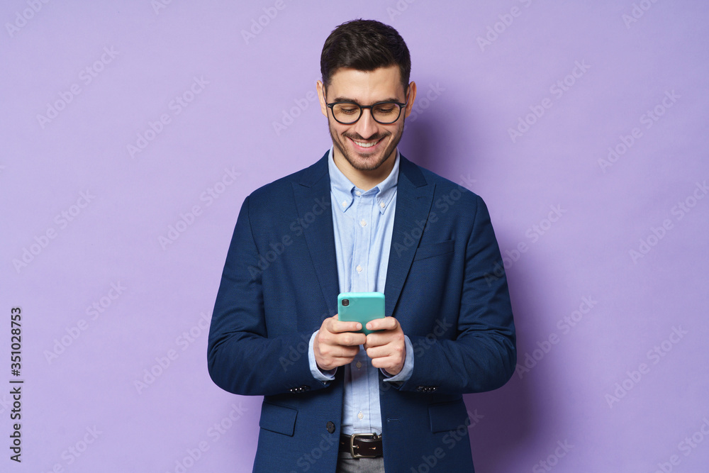 Young man wearing formal clothes, looking at phone screen, exchanging messages with friends or colleagues, smiling happily, isolated on purple background Young man wearing formal clothes, looking at phone screen, exchanging messages with friends or colleagues, smiling happily, isolated on purple background