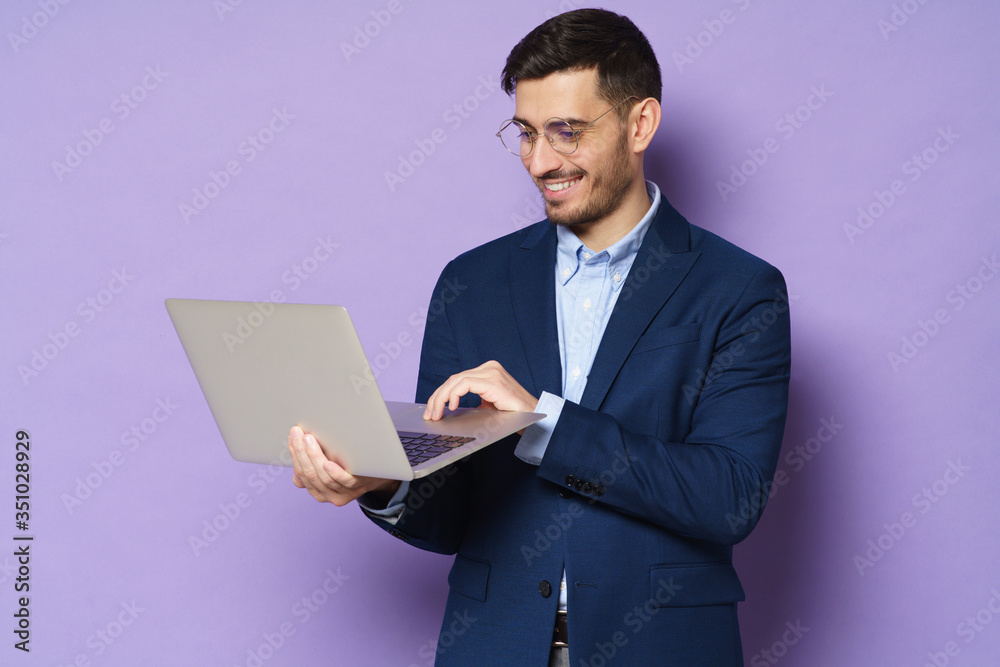 Young businmessman wearing trendy smart casual suit, standing with open laptop in hands, smiling at content on screen, isolated on purple background
