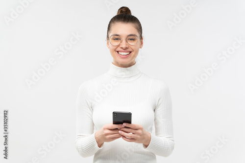 Female looking at camera with smile, isolated on gray background, holding cellphone in front of her, communicating with friends, feeling relaxed and happy © Damir Khabirov
