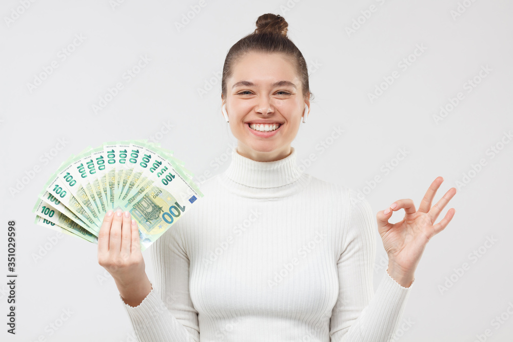 Pretty girl dressed in white turtleneck smiling happily, showing euro banknotes in cash and ok sign as if satisfied with prize, isolated on gray background