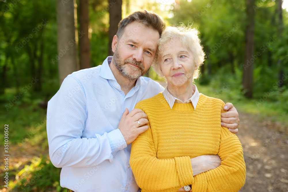 Loving adult son tenderly embracing his joyful elderly mother during walking at summer park. Mother's day holiday. Loving adult son tenderly embracing his joyful elderly mother during walking at summer park. Mother's day holiday.