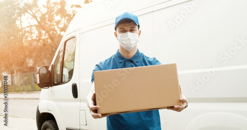 Portrait of young courier in mask standing near delivery car and holding carton box © VAKSMANV