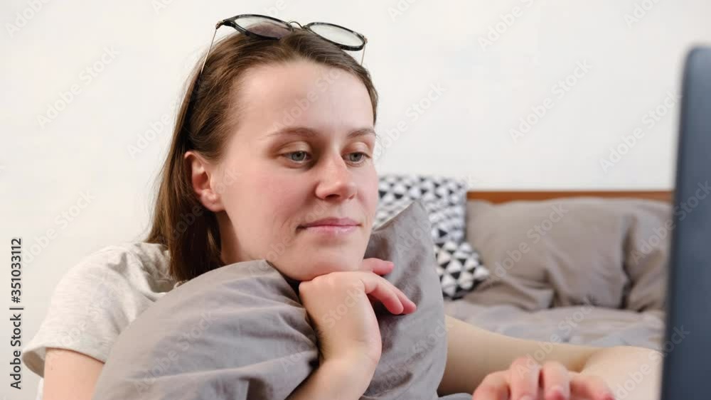 Cheerful millennial woman user having fun surfing internet, watching social media videos online. Happy young woman in eyeglasses laughing using modern laptop sitting on bed at home. Cheerful millennial woman user having fun surfing internet, watching social media videos online. Happy young woman in eyeglasses laughing using modern laptop sitting on bed at home.