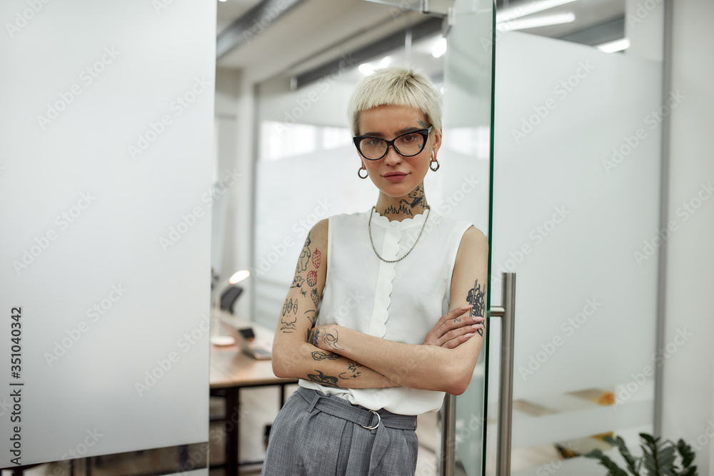 Successful business lady. Young beautiful blonde tattooed businesswoman with short haircut leaning on the glass door, keeping arms crossed and looking at camera while standing in modern office Successful business lady. Young beautiful blonde tattooed businesswoman with short haircut leaning on the glass door, keeping arms crossed and looking at camera while standing in modern office