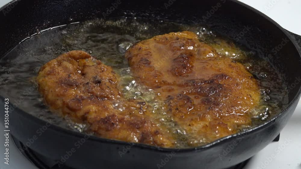 Two pieces of breaded chicken are shown being fried in a cast iron pan in a closeup view. Two pieces of breaded chicken are shown being fried in a cast iron pan in a closeup view.