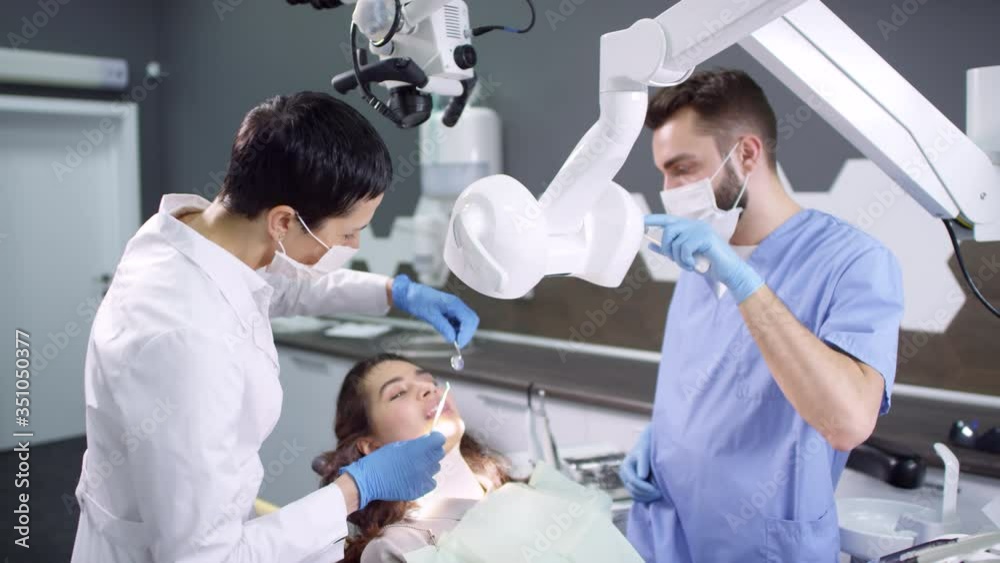 Medium shot of male dental assistant helping female dentist examining teeth of patient sitting on dental chair Medium shot of male dental assistant helping female dentist examining teeth of patient sitting on dental chair