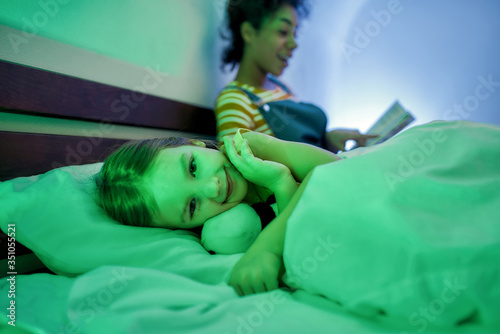 A warm surrounding. African american woman baby sitter reading book to cute little girl in the evening. Nanny holding book while kid listening to fairy tale, falling asleep