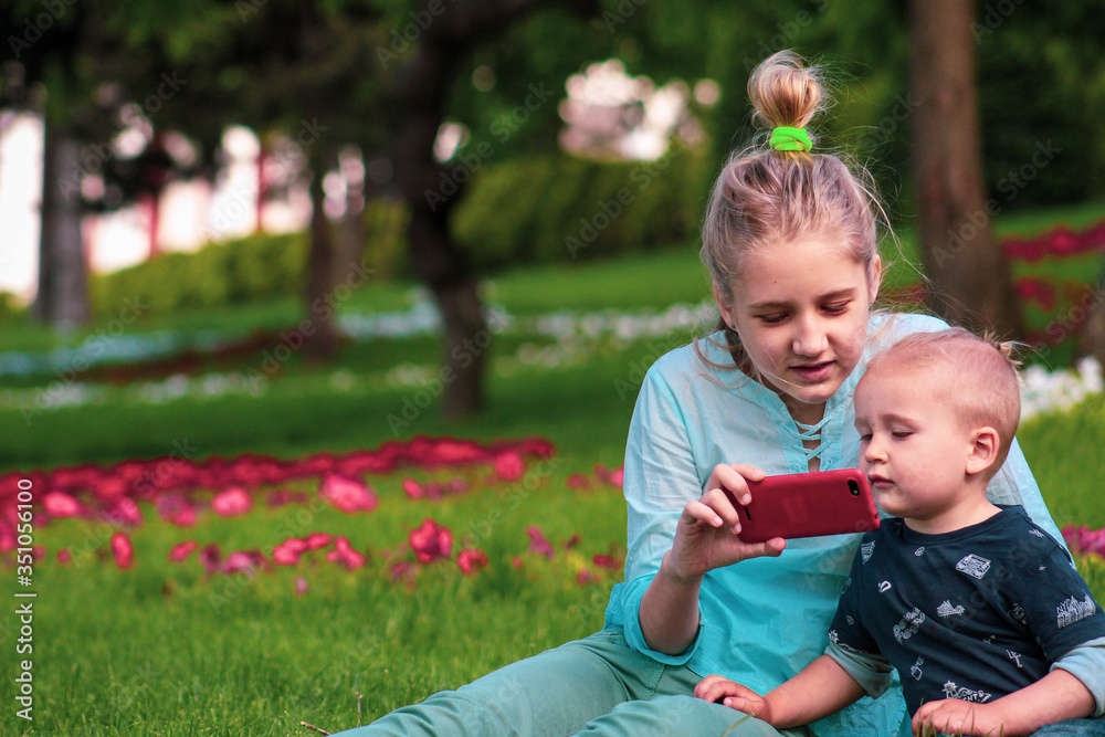 Blond older sister and younger brother of European appearance are sitting on the green grass and watching a video on the phone in the spring in the park. Blond older sister and younger brother of European appearance are sitting on the green grass and watching a video on the phone in the spring in the park.