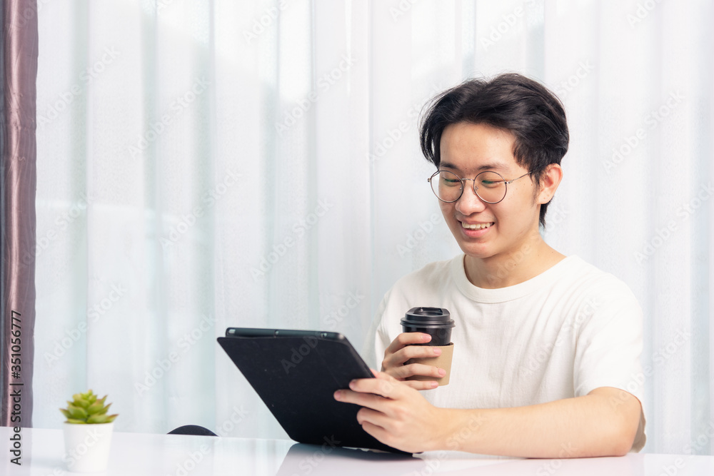 Happy Asian young business handsome man work from home office wear glasses, t-shirt comfortable he smiling and using a black modern smart digital tablet computer to read email or learning on desk Happy Asian young business handsome man work from home office wear glasses, t-shirt comfortable he smiling and using a black modern smart digital tablet computer to read email or learning on desk