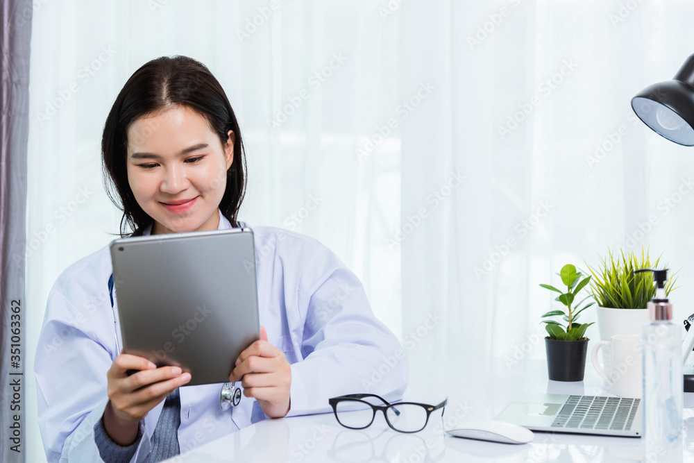Asian young beautiful woman doctor on desk front laptop computer she holding modern smart digital tablet on hand during quarantines disease coronavirus or COVID-19 at hospital office Asian young beautiful woman doctor on desk front laptop computer she holding modern smart digital tablet on hand during quarantines disease coronavirus or COVID-19 at hospital office