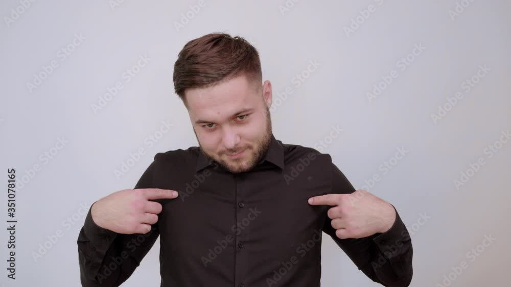 Young Bearded Dark Haired Man In Black Stylish Shirt On White Background, Conceived Male Shows Forefingers On Himself And Thinks. The Concept Is Focused On Smart People. Young Bearded Dark Haired Man In Black Stylish Shirt On White Background, Conceived Male Shows Forefingers On Himself And Thinks. The Concept Is Focused On Smart People.
