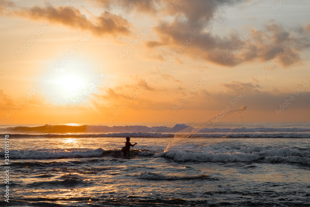 A fisherman throws a net into the sea at dawn. Beautiful sunrise at Pantai Pabean Ketewel Beach on the east coast of Bali.