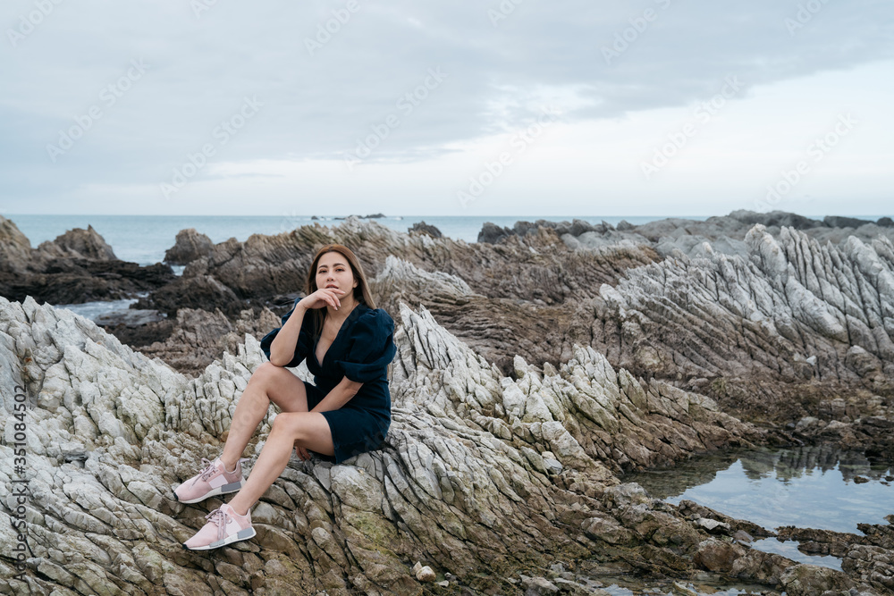 Portrait of young asian woman on stones near sea. Beautiful girl is resting on coast, enjoying outdoor recreation at Kaikoura, New zealand.