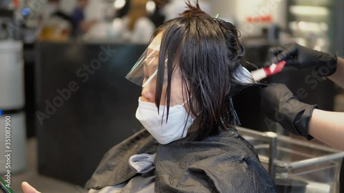 Hairdresser in gloves with Brush In Hand Applying Mask On short Healthy Hair. Beautiful Asian Girl wearing a protective mask at Hair Salon. Spreading Conditioner Along with Wet Hair. Health And Beauty photo