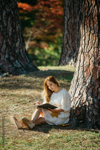 Beautiful asian woman in white dress sit under the tree  writing and thinking in the park with autumn leaves. © tonklafoto