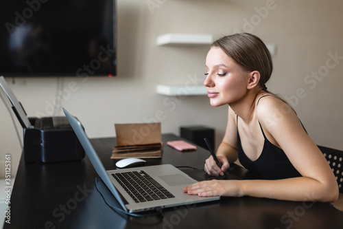Beautiful girl freelancer businessman works with a graphics tablet in an image editor and looks at the monitor. Work at home during an epidemic. Self-isolation.