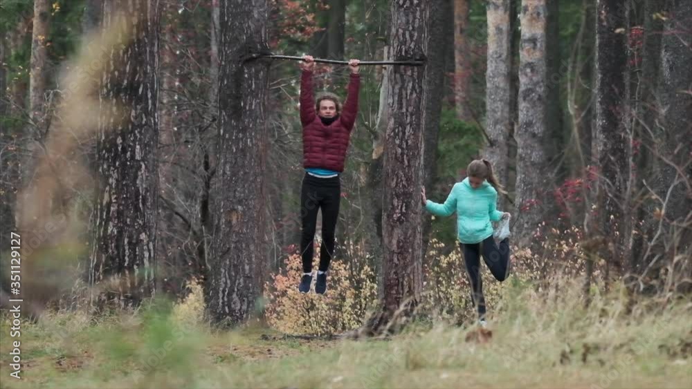 Young couple training. Man and woman work out in the autumn forest. Man pulls up on the bar, woman stretches Young couple training. Man and woman work out in the autumn forest. Man pulls up on the bar, woman stretches