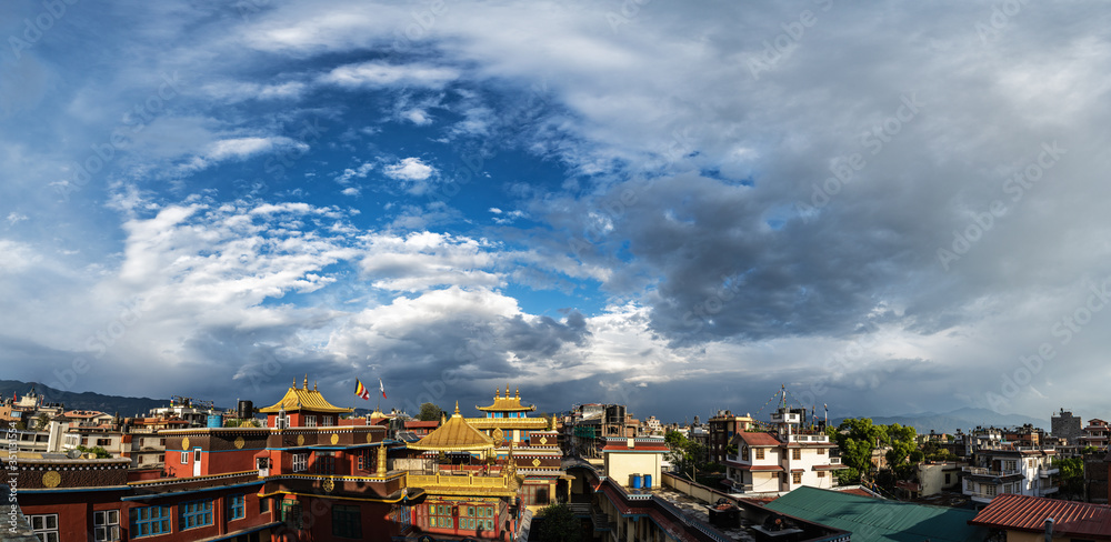 Panoramic view of the roofs of houses and temples of the Kathmandu Valley, gilded by sunlight. The clouds in the sky above the houses are twisted into a spiral.