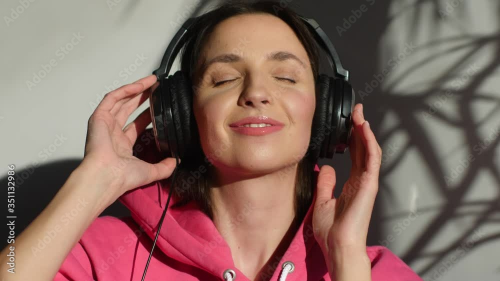 brunette girl in a pink hoodie sits at home on the sofa and listens to music in large headphones, in a sharp light against a white wall brunette girl in a pink hoodie sits at home on the sofa and listens to music in large headphones, in a sharp light against a white wall