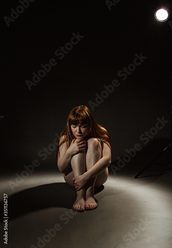 A single naked girl is sitting with her arms around her knees on a black background