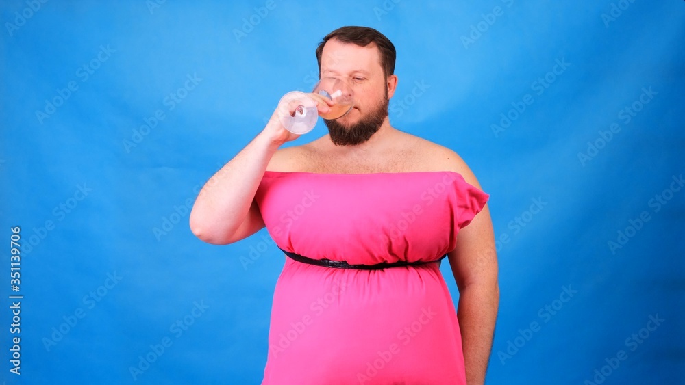 Funny bearded guy in a pink dress made of pillows drinks wine from a glass on a blue background. Crazy quarantine. Fashion 2020. Put on a pillow. Challenge 2020 due to house isolation Funny bearded guy in a pink dress made of pillows drinks wine from a glass on a blue background. Crazy quarantine. Fashion 2020. Put on a pillow. Challenge 2020 due to house isolation