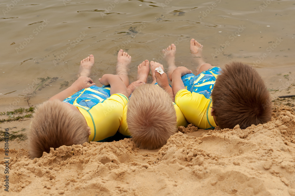 Funny kids in bathing suits sit together on sand with their legs in water. boys are happy on holiday in village together. Summer day, river, swimming in the water. Close-up legs and feet Funny kids in bathing suits sit together on sand with their legs in water. boys are happy on holiday in village together. Summer day, river, swimming in the water. Close-up legs and feet