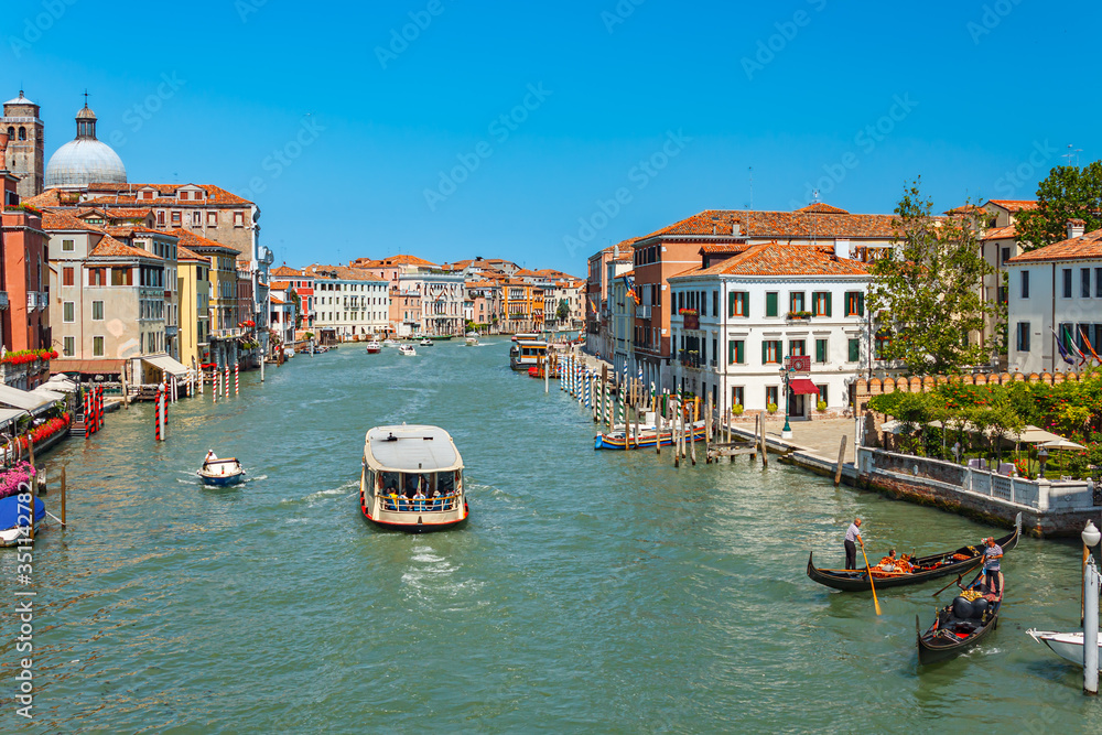 Tourist boat sailing along the Grand Canal with emerald green water past ancient houses with tiled roofs, brightly lit by the sun. Venice, Italy.