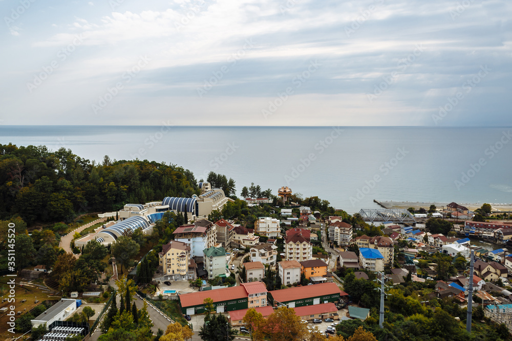 View of a small resort town near the city of Sochi. The photo shows the Black Sea, small hotels, residential buildings, green trees and blue sky.