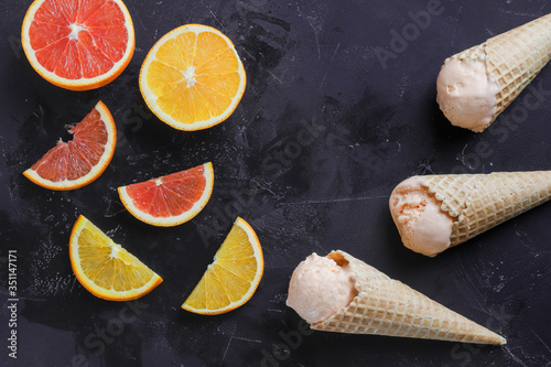orange ice cream in a waffle cone on a dark surface, in the frame whole oranges, orange slices, orange rings, top view.