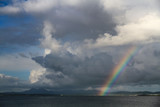 Tiefhängende Gewitterwolken mit Regenbogen über einer Meeresbucht mit sonnenbeleuchtetem Fischerort am anderen Ufer