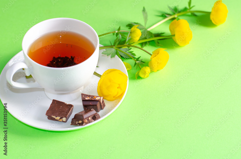 A Cup of black tea, pieces of chocolate and yellow flowers of the Trollius europaeus on a green background. Sunny, bright, shadows fall from the window. Chocolate day and morning tea ceremony. A Cup of black tea, pieces of chocolate and yellow flowers of the Trollius europaeus on a green background. Sunny, bright, shadows fall from the window. Chocolate day and morning tea ceremony.