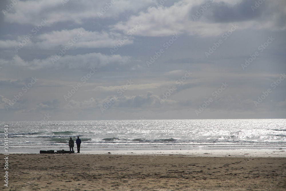 Zwei Personen stehen weit entfernt an einem Strand bei grauen Wolken und sonnenglitzerndem Wasser