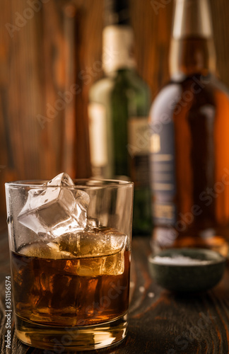 Fototapeta Naklejka Na Ścianę i Meble -  A glass of whiskey with ice, in the background are bottles on a wooden table of a bar counter, shallow depth of field, selective focus. The concept of alcoholic drinks in a roadside bar.