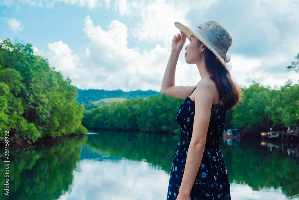 Asian women standing, wearing a hat and admiring the River view with mangrove forest. And the clear skies with beautiful clouds. Suitable for tourism, recreation and relax