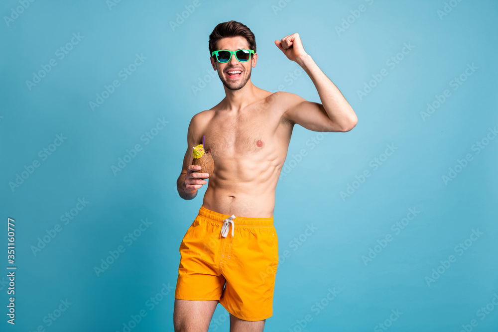 Photo of cheerful positive handsome man making fist raising hand to say yeah smiling toothily holding cup of coconut with beverage inside isolated pastel blue color background